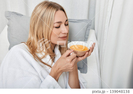 Pretty caucasian woman in white bathrobe drinking hot lemon tea enjoying spa procedures in wellness center Pretty caucasian woman in white bathrobe drinking hot lemon tea enjoying spa procedures in wellness center 139048138