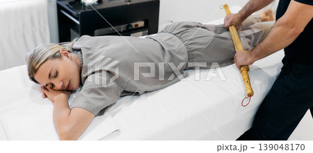 Caucasian woman receiving anti-stress massage with bamboo sticks at massage center. Bamboo brooms in masseur hands during massage Caucasian woman receiving anti-stress massage with bamboo sticks at massage center. Bamboo brooms in masseur hands during massage 139048170