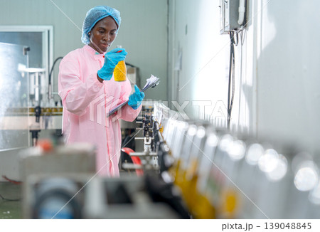 Front view of African American woman factory worker who hold document file and bottle with yellow liquid to check quality of product in line productivity in drinking industrial factory workplace. Front view of African American woman factory worker who hold document file and bottle with yellow liquid to check quality of product in line productivity in drinking industrial factory workplace. 139048845