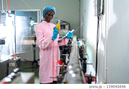 Wide shot of African American woman factory worker who hold document file and bottle with yellow liquid to check quality of product in line productivity in drinking factory workplace. Wide shot of African American woman factory worker who hold document file and bottle with yellow liquid to check quality of product in line productivity in drinking factory workplace. 139048846