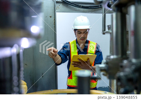 Asian factory technician or engineer hold document file and point to part of containing machine to check the quality of the line product and function of machine awith standard control for good Asian factory technician or engineer hold document file and point to part of containing machine to check the quality of the line product and function of machine awith standard control for good 139048855