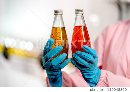 Close up hands with blue glove of factory worker hold the bottle with red and yellow liquid to compare quality of the product in drinking factory workplace. Close up hands with blue glove of factory worker hold the bottle with red and yellow liquid to compare quality of the product in drinking factory workplace. 139048860