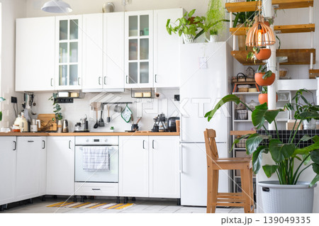Modern kitchen interior, lots of houseplants, window light, white cabinets, eco-style, potted greenery, wooden modular stair detail, spacious and fresh look. Modern kitchen interior, lots of houseplants, window light, white cabinets, eco-style, potted greenery, wooden modular stair detail, spacious and fresh look. 139049335