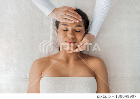 Beautiful black woman lying on spa bed while therapist gently massages head Beautiful black woman lying on spa bed while therapist gently massages head 139049907