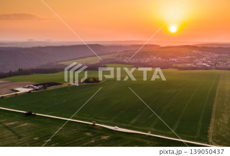 Aerial view of agriculture land during beautiful sunset. Fields and meadow with the small village in far. Vibrant soft colors at magic dusk. The asphalt road leading between fields with tree on side Aerial view of agriculture land during beautiful sunset. Fields and meadow with the small village in far. Vibrant soft colors at magic dusk. The asphalt road leading between fields with tree on side 139050437