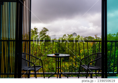 View from hotel room window with balcony to tropical forest during the storm with rain. Big shadow clouds and rainy weather. Two chairs on the side with simple table. View from hotel room window with balcony to tropical forest during the storm with rain. Big shadow clouds and rainy weather. Two chairs on the side with simple table. 139050619