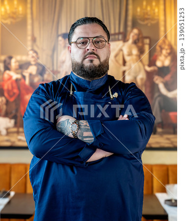 Chef portrait in restaurant, close cropped detail showing folded arms and tattoos, navy jacket texture and calm intensity highlight 139051253