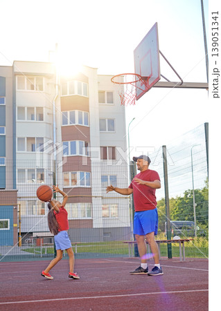 The girl makes a powerful upward throw of the ball. The father trains the child in the yard on the court playing basketball The girl makes a powerful upward throw of the ball. The father trains the child in the yard on the court playing basketball 139051341