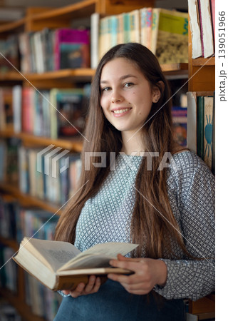 Young smiling girl reading a look. Student with an open book standing near shelves in library, choosing literature for reading Young smiling girl reading a look. Student with an open book standing near shelves in library, choosing literature for reading 139051966