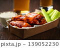 Buffalo BBQ chicken wings with blue cheese sauce and beer with of celery sticks in a paper container on a wooden table.Buffalo chicken wings in a disposable tray and beer on a wooden table 139052230