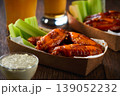Buffalo BBQ chicken wings with blue cheese sauce and beer with of celery sticks in a paper container on a wooden table.Buffalo chicken wings in a disposable tray and beer on a wooden table 139052232
