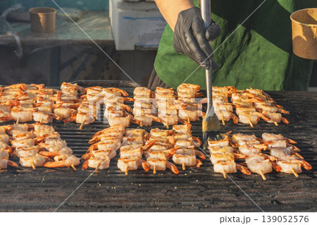 Shrimp skewers grilling on barbecue grill, chef brushes sauce, close view of seafood cooking process with rows of shrimp turning golden on hot surface. Shrimp grilling, street food barbecue Shrimp skewers grilling on barbecue grill, chef brushes sauce, close view of seafood cooking process with rows of shrimp turning golden on hot surface. Shrimp grilling, street food barbecue 139052576