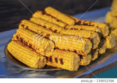 Grilled corn cobs stacked on metal tray with charred marks and bright yellow kernels prepared in outdoor street food market. Street food, grilled corn, barbecue cooking, fresh vegetable snack Grilled corn cobs stacked on metal tray with charred marks and bright yellow kernels prepared in outdoor street food market. Street food, grilled corn, barbecue cooking, fresh vegetable snack 139052586