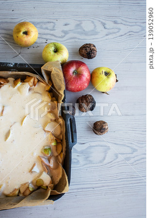 Apple pie batter poured over sliced apples in a baking pan, with rustic background, alongside fresh apples and walnuts. 139052960