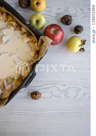 Apple pie batter poured over sliced apples in a baking pan, with rustic background, alongside fresh apples and walnuts. 139052966