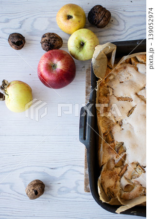 Homemade apple cake with a golden crust in a baking tray, placed on a rustic white wooden table with fresh apples and walnuts 139052974