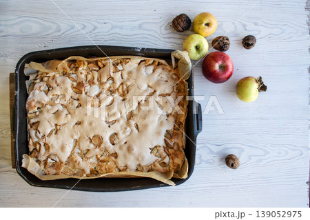 Homemade apple cake with a golden crust in a baking tray, placed on a rustic white wooden table with fresh apples and walnuts Homemade apple cake with a golden crust in a baking tray, placed on a rustic white wooden table with fresh apples and walnuts 139052975