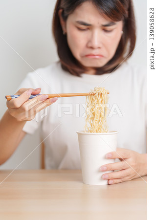 Bored young Asian woman eating instant noodles at home. Exhausted female looking unhappy while eating cup ramen. quick meal after work, burnout and unhealthy eating habit, lifestyle and cheap meal Bored young Asian woman eating instant noodles at home. Exhausted female looking unhappy while eating cup ramen. quick meal after work, burnout and unhealthy eating habit, lifestyle and cheap meal 139058628