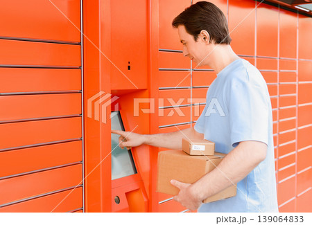 Man with cardboard boxes uses the touch screen of an automated orange parcel locker to pick up or drop off packages. Convenient e-commerce and self-service delivery tech. Man with cardboard boxes uses the touch screen of an automated orange parcel locker to pick up or drop off packages. Convenient e-commerce and self-service delivery tech. 139064833