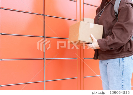 A woman holds cardboard boxes in front of an automated orange mailbox. Concept for self-service delivery, online order returns, and delivery logistics. A woman holds cardboard boxes in front of an automated orange mailbox. Concept for self-service delivery, online order returns, and delivery logistics. 139064836