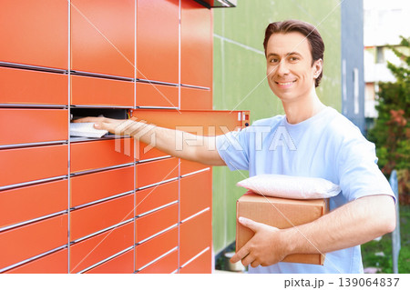 Happy young man smiling at the camera while picking up or sending packages at an automated orange parcel locker outdoors. Convenient e-commerce and self-service delivery concept. 139064837