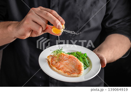 Chef squeezing lemon over grilled fish fillet with vegetables in restaurant kitchen close up 139064919