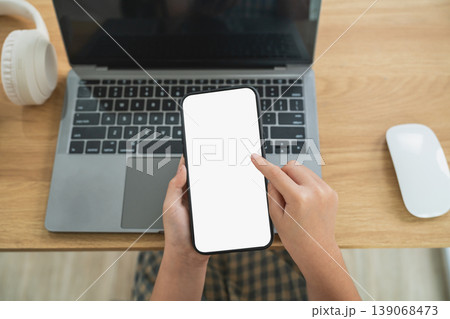 Person Holding Smartphone with Blank Screen Above Laptop on Wooden Table with Headphones and Mouse in Modern Workspace Setting for Technology Use Person Holding Smartphone with Blank Screen Above Laptop on Wooden Table with Headphones and Mouse in Modern Workspace Setting for Technology Use 139068473