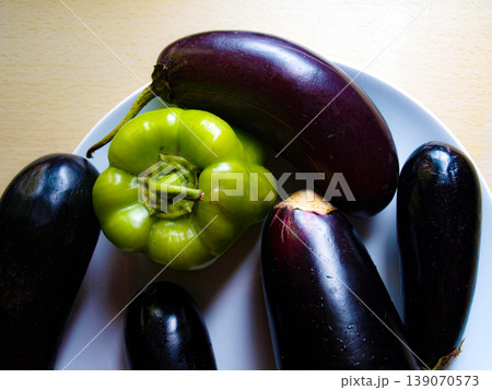 Green bell pepper and purple eggplant on a white plate. Still life composition on a wooden table. Contrast of vibrant vegetables, porcelain and rustic wood texture in natural light. 139070573