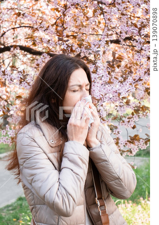 Vertical shot of a woman blowing her nose outdoors. Spring allergy symptom, hay fever relief, seasonal cold. 139070898