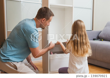 A father and daughter work together to place a white drawer into their newly assembled shelf in a bright living room with a gray sofa in the background A father and daughter work together to place a white drawer into their newly assembled shelf in a bright living room with a gray sofa in the background 139070921