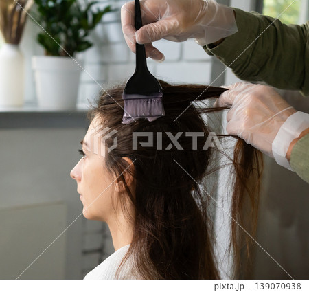 gloved hand applies dye or treatment to a woman's hair roots with brush in bright room. The concept of home care in beauty salon gloved hand applies dye or treatment to a woman's hair roots with brush in bright room. The concept of home care in beauty salon 139070938