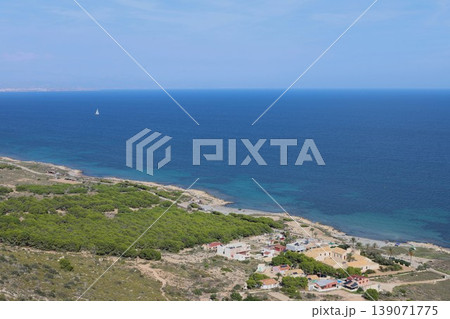 Coastal landscape of Spain showing the deep blue sea, a distant sailboat, and green vegetation next to a small village Coastal landscape of Spain showing the deep blue sea, a distant sailboat, and green vegetation next to a small village 139071775