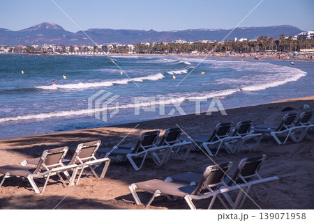 Beach scene with empty lounge chairs on sandy shore, gentle waves lapping at the coast and distant mountains under clear blue sky Beach scene with empty lounge chairs on sandy shore, gentle waves lapping at the coast and distant mountains under clear blue sky 139071958