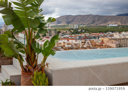 Rooftop infinity pool with tropical plants overlooking a cityscape and mountains under a cloudy sky in a Mediterranean region Rooftop infinity pool with tropical plants overlooking a cityscape and mountains under a cloudy sky in a Mediterranean region 139071959