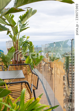 Rooftop terrace with wicker chairs and tropical plants overlooking a city skyline under a cloudy sky 139071969