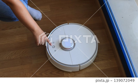 Person using a robotic vacuum cleaner on wooden floor, pointing at control buttons near a window with natural light streaming in Person using a robotic vacuum cleaner on wooden floor, pointing at control buttons near a window with natural light streaming in 139072049