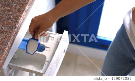 Person adding detergent to washing machine drawer in a bright laundry room with blue window and granite countertop 139072083
