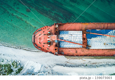 An aerial view shows a large, abandoned red cargo ship beached on a white pebble coastline. 139073003