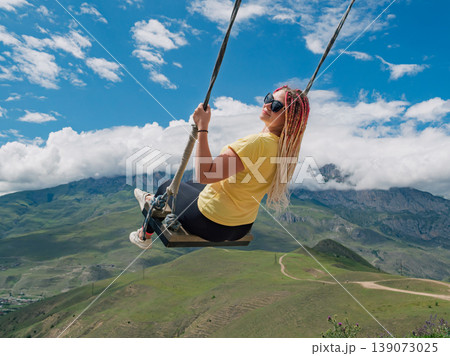 Woman enjoying mountain swing ride overlooking the valley with vast green landscapes of Caucasus Mountains and blue sky Woman enjoying mountain swing ride overlooking the valley with vast green landscapes of Caucasus Mountains and blue sky 139073025