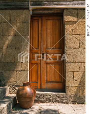 Brown wooden double door framed by rough hewn stone walls. A large decorative clay pot rests on sunlit stone steps leading to the entrance. 139073032