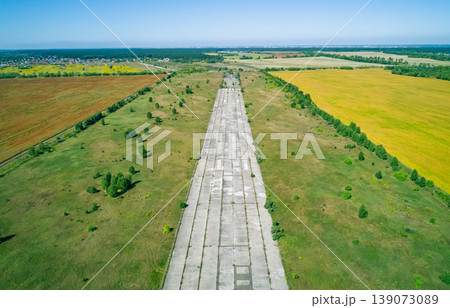 Aerial view of a long, cracked concrete runway at an abandoned airfield, flanked by green fields and dense trees. Aerial view of a long, cracked concrete runway at an abandoned airfield, flanked by green fields and dense trees. 139073089