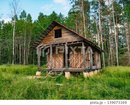 Weathered wooden cabin stands abandoned in a lush green forest clearing under a bright summer sky 139073169