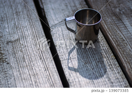 Stainless steel mug on rustic wooden table outdoors, reflecting sunlight. Metal coffee cup. Minimalism, travel essentials, camping, hiking, sustainable living in nature. Selective focus Stainless steel mug on rustic wooden table outdoors, reflecting sunlight. Metal coffee cup. Minimalism, travel essentials, camping, hiking, sustainable living in nature. Selective focus 139073385