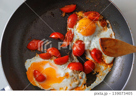 Overhead view of two fried eggs cooked with cherry tomato pieces in a dark frying pan, with a wooden spatula, Simple homemade breakfast. Cooking scene Overhead view of two fried eggs cooked with cherry tomato pieces in a dark frying pan, with a wooden spatula, Simple homemade breakfast. Cooking scene 139073405