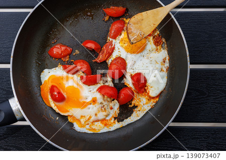 Overhead view of fried eggs cooked with cherry tomatoes in a frying pan on a black table with a wooden spatula. Simple home cooking and breakfast concept with copy space 139073407