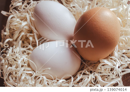 Three fresh chicken eggs laying in a nest of wood shavings, including two white eggs and one brown egg 139074014