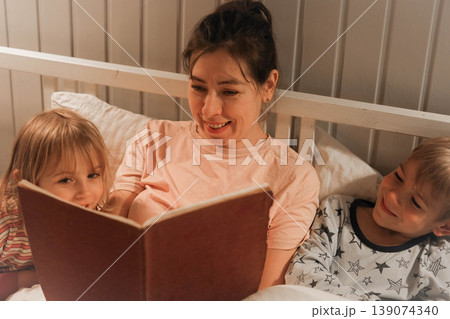 Family bedtime stories. A mother reads a story to her children, wrapped in a blanket. Children listen to soothing stories. A cozy family scene before bed in a softly lit bedroom. Copy space. Banner 139074340