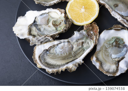 Khasan oysters, live, fresh, opened. Served with classic French Mignonette sauce and half a lemon. Blue plate, black background. Selective focus, close-up. 139074538