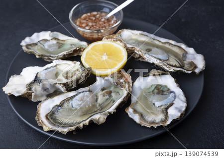 Khasan oysters, live, fresh, opened. Served with classic French Mignonette sauce and half a lemon. Blue plate, black background. Selective focus, close-up. 139074539