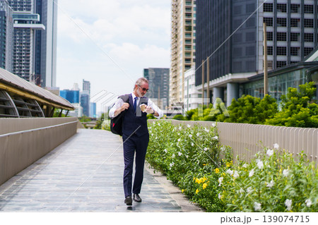 Professional senior Caucasian businessman in navy vest and tie, holding a coffee cup and carrying a backpack. He is looking up at urban skyscrapers in modern business district. Portrait of senior male Professional senior Caucasian businessman in navy vest and tie, holding a coffee cup and carrying a backpack. He is looking up at urban skyscrapers in modern business district. Portrait of senior male 139074715